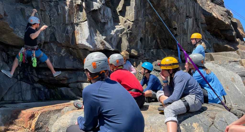 A group of young people wearing safety gear watch a person during a rock climbing exercise. 